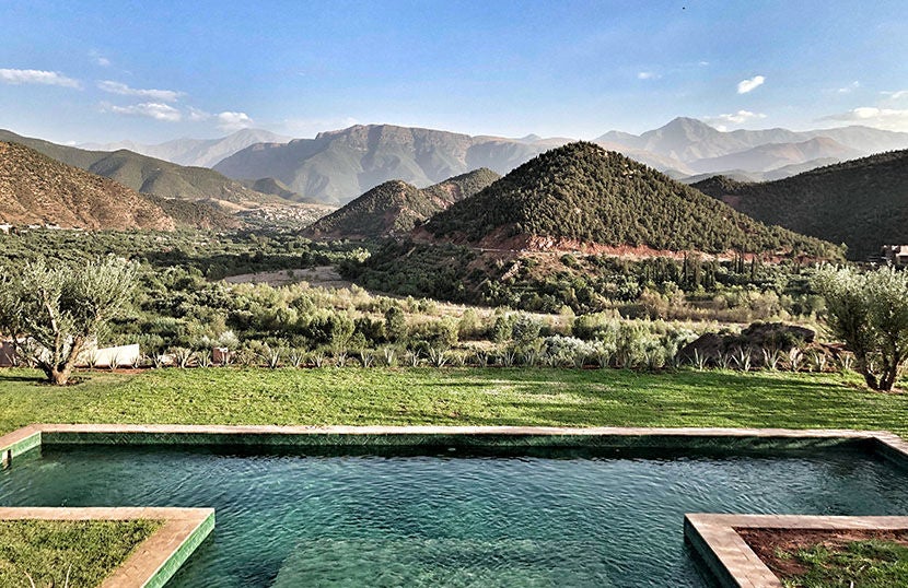 Elegant Moroccan hotel with terracotta walls nestled in Atlas Mountains, featuring infinity pool overlooking lush valley at sunset