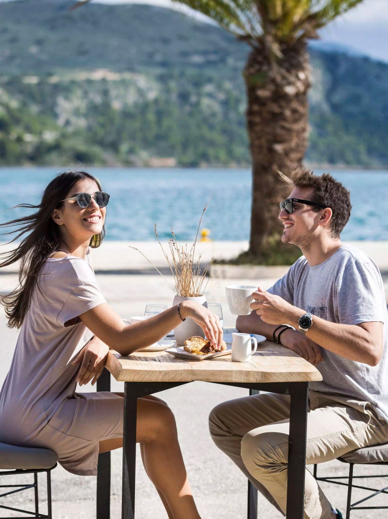 Luxurious seaside hotel with white Mediterranean architecture, blue-domed roofs, nestled against dramatic Greek coastline with azure Ionian Sea backdrop