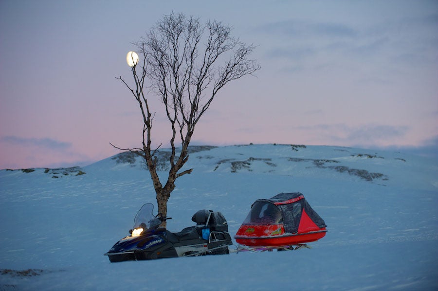 Snowmobile riders traverse pristine snowy terrain in Norway, surrounded by majestic mountain peaks and untouched wilderness at sunset