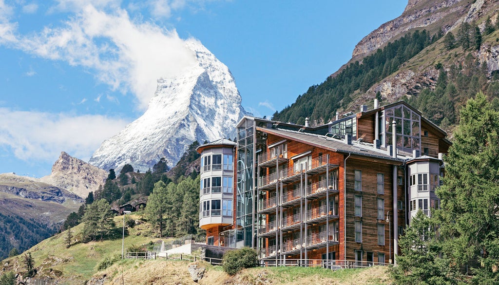 Modern Alpine hotel nestled in snowy mountains, featuring a sleek glass-walled restaurant terrace with panoramic views of Zermatt valley