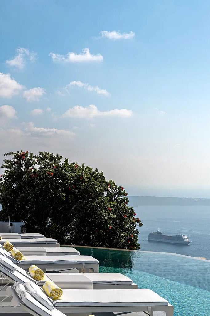 Elegant white stone villa with infinity pool overlooking Aegean Sea, nestled into Santorini cliffside at sunset with blue domed churches