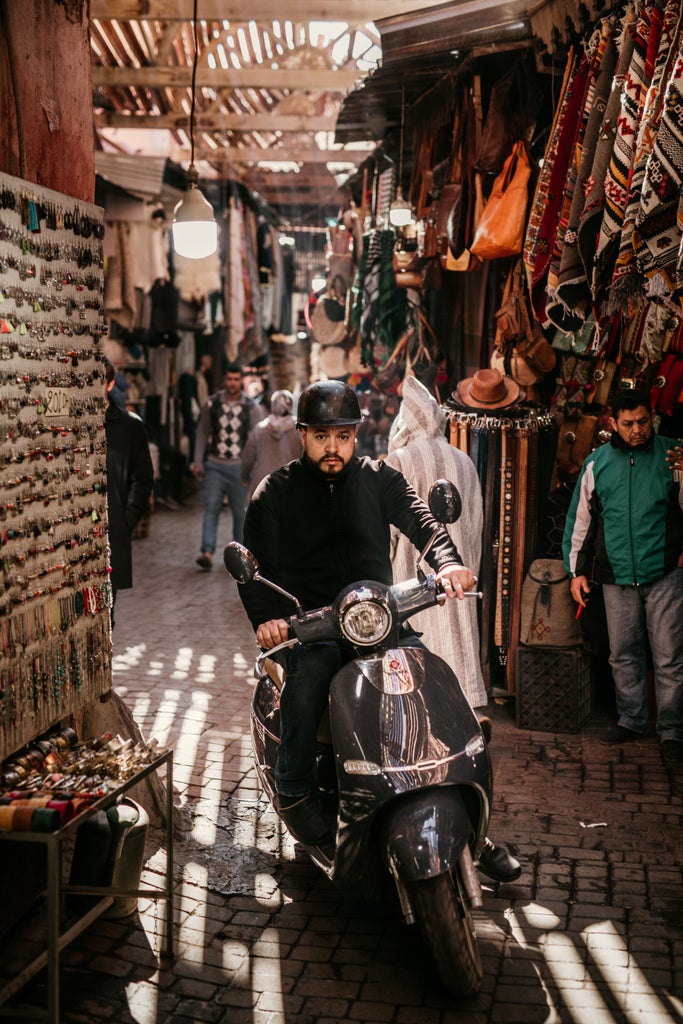 Tourists riding vintage motorcycles through narrow, winding streets of Marrakech's medina, passing ornate riads and bustling souks