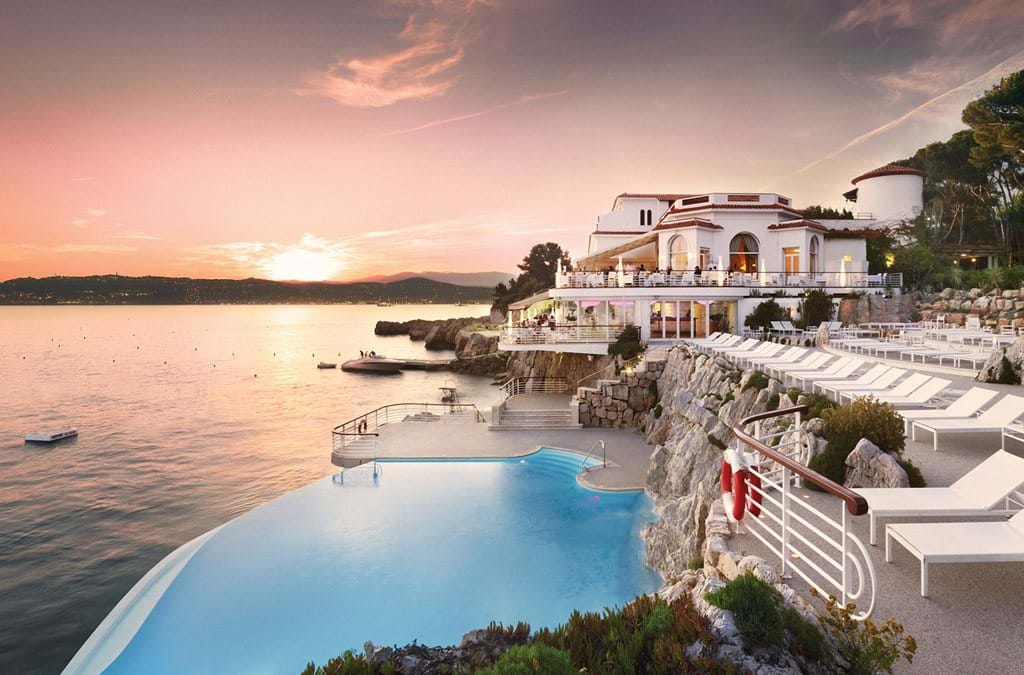 Elegant seaside luxury hotel with cream facade, ornate balconies and manicured gardens overlooking Mediterranean Sea at sunset