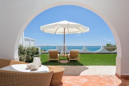 Elegant hotel suite with ocean-view balcony, featuring cream decor, plush seating area, and modern chandelier in soft natural light