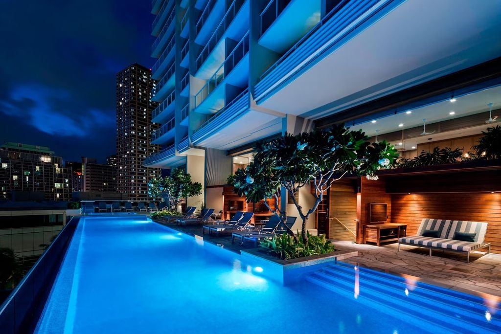 Luxurious oceanfront high-rise hotel with white balconies, palm trees and tropical gardens, set against Waikiki's turquoise waters