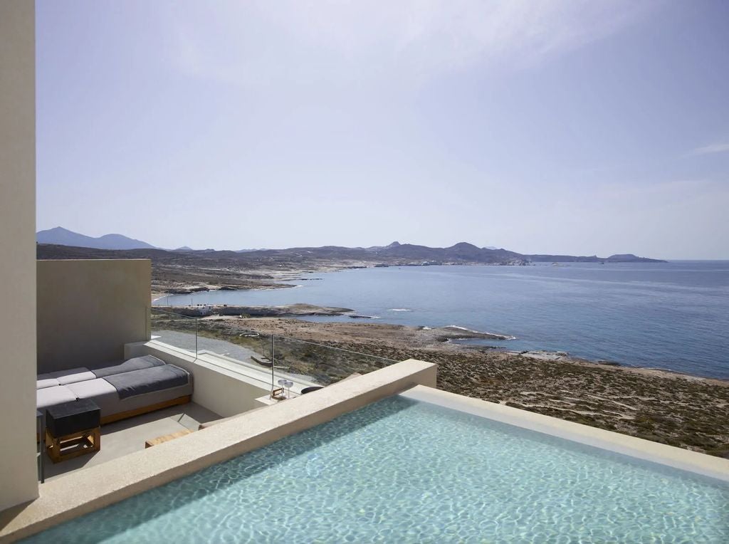 Elegant white suites with infinity pools cascade down cliffside overlooking Aegean Sea, stone pathways weave through Cycladic architecture