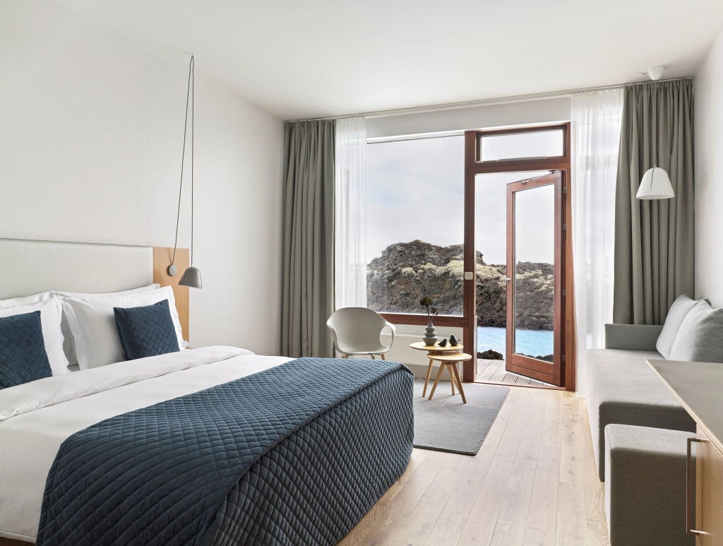 Minimalist Icelandic hotel room with panoramic windows, natural volcanic stone walls, crisp white linens, and modern Nordic design elements overlooking dramatic landscape