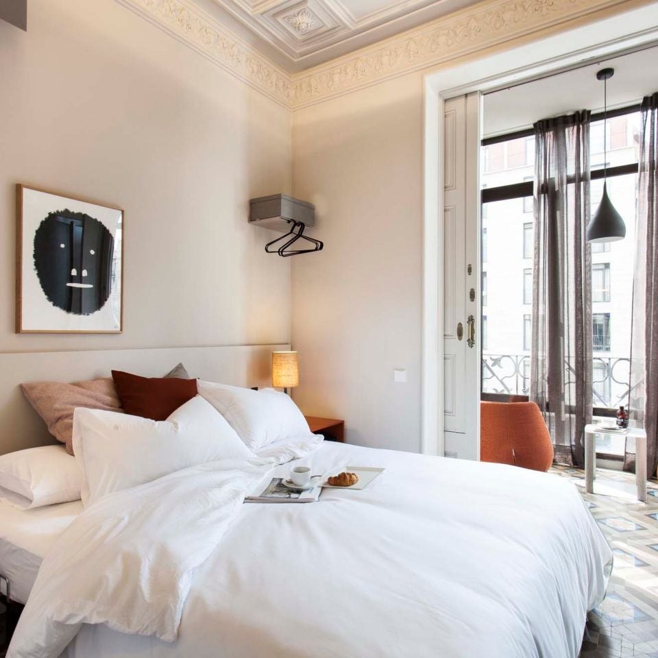 Elegant modern hotel room with minimalist design, white linens, wooden floor, and panoramic window overlooking urban Barcelona skyline at sunset