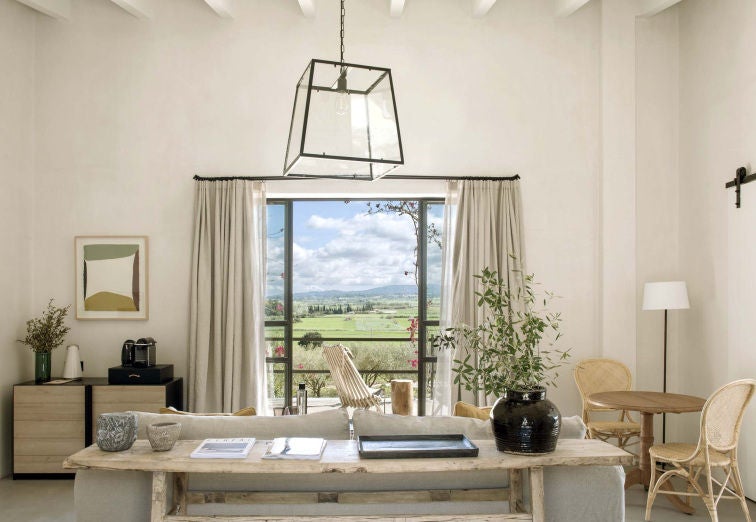 Elegant rustic-luxury hotel room with stone walls, high wooden beams, cream linens, and panoramic windows overlooking Spanish countryside