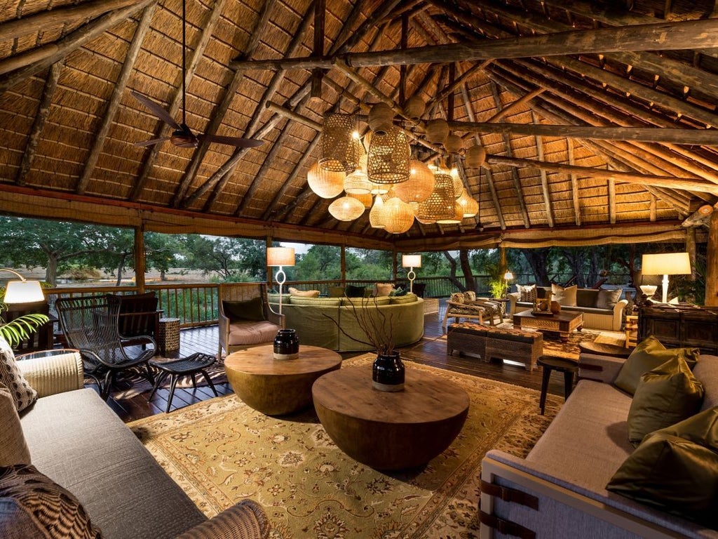 Luxury safari lodge overlooking a tranquil river, featuring a stone deck, infinity pool, and thatched roof amid African bush landscape