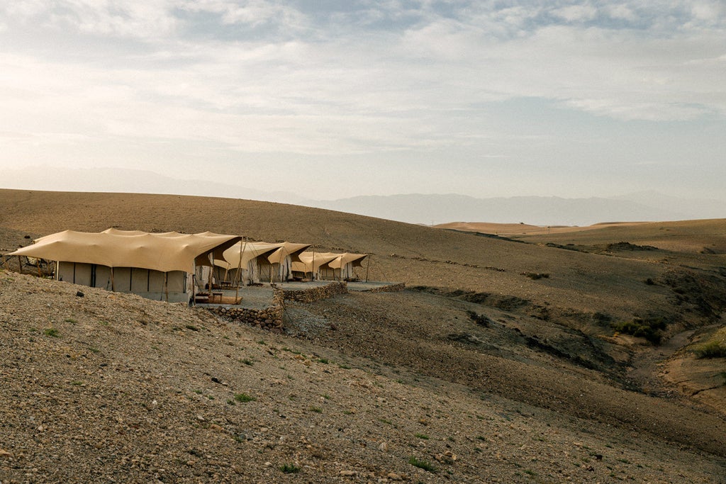 Luxurious desert tent featuring king bed, woven rugs, modern furnishings, and panoramic views of Agafay's rocky terrain at sunset