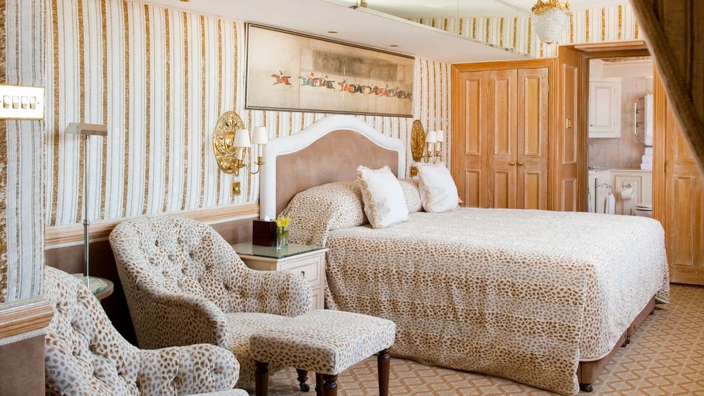 Elegant deluxe hotel room with plush king bed, sophisticated cream and gold color palette, ornate decorative details, and refined British luxury styling