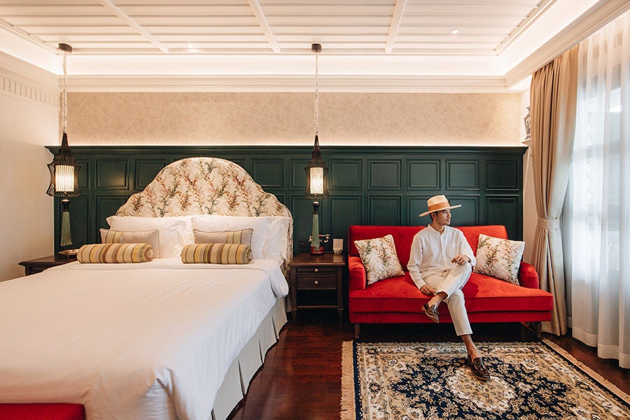 Elegant corner suite with traditional Lanna-style decor, featuring a plush king bed, wooden furnishings, and floor-to-ceiling windows overlooking nature