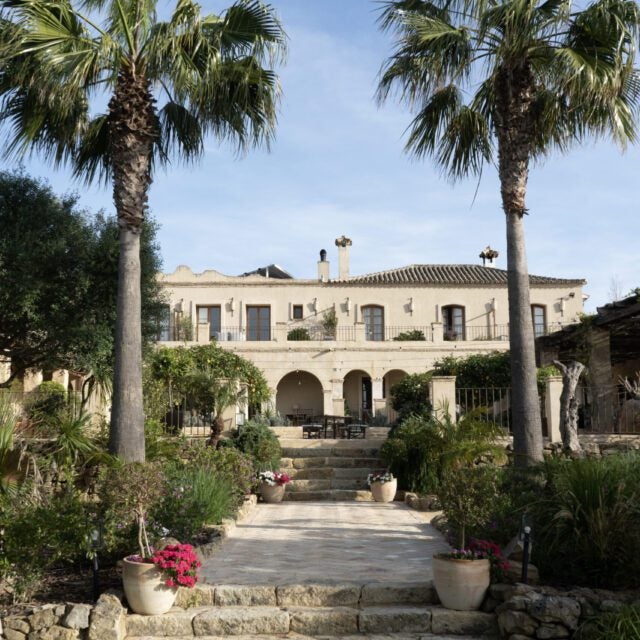 Luxurious boutique hotel with whitewashed Mediterranean facade, terracotta roof tiles, and elegant arched windows amid lush garden landscaping