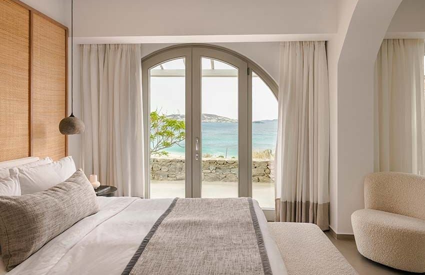 Elegant white-washed Cycladic-style deluxe hotel room with sweeping blue sea view, minimalist design, and private balcony overlooking Mykonos coastline