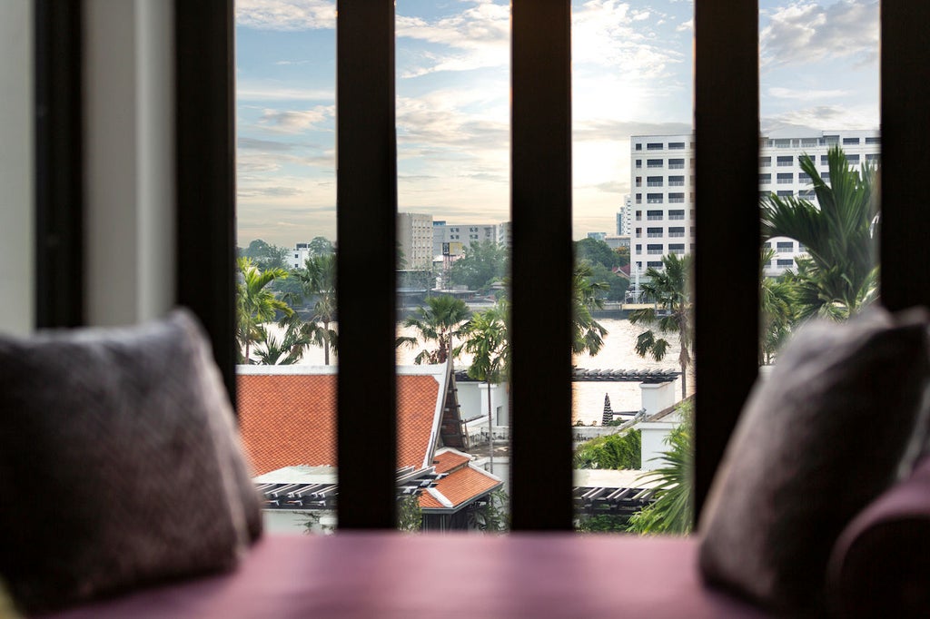 Elegant suite with river views featuring modern Thai decor, king bed, spacious sitting area and floor-to-ceiling windows in neutral tones