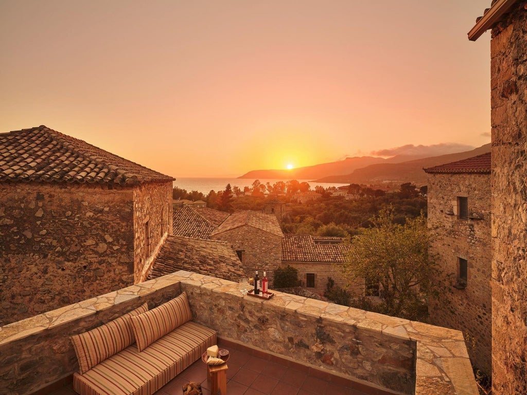 Stone-walled Mediterranean villa with terracotta roof nestled in lush Greek landscape, featuring elegant outdoor seating and panoramic Aegean Sea view