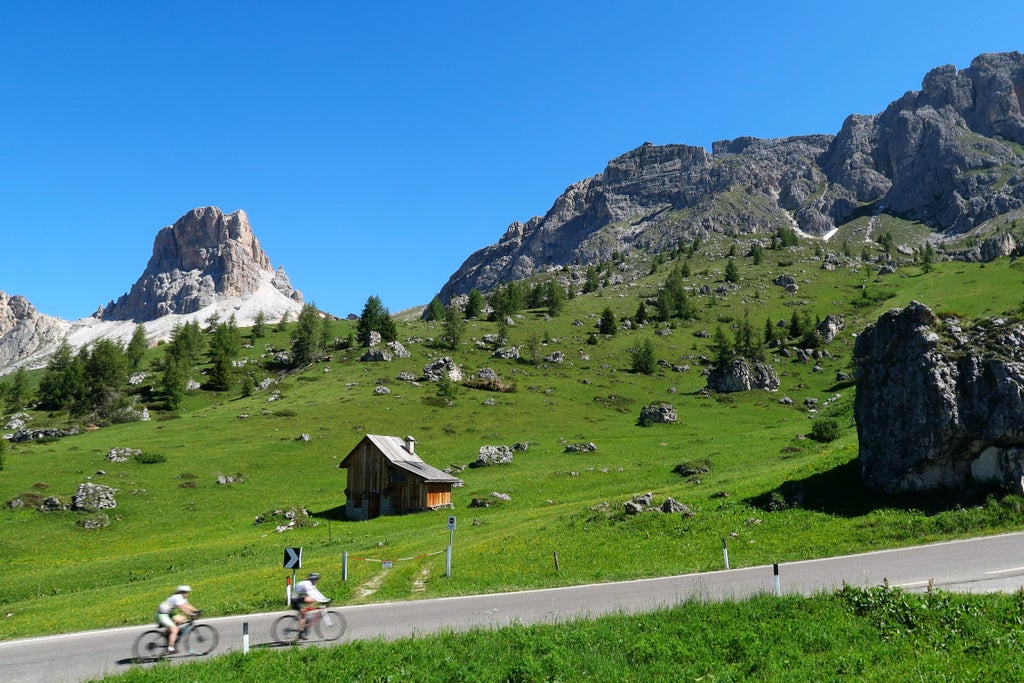 Snow-capped Dolomite mountain peaks rise majestically over alpine meadow, with a rustic wooden cabin nestled beneath dramatic rocky cliffs