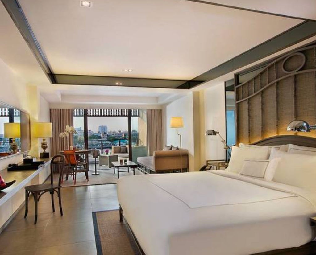 Modern hotel room with plush king bed, white linens, dark wood furnishings, floor-to-ceiling windows and elegant Thai-inspired decor