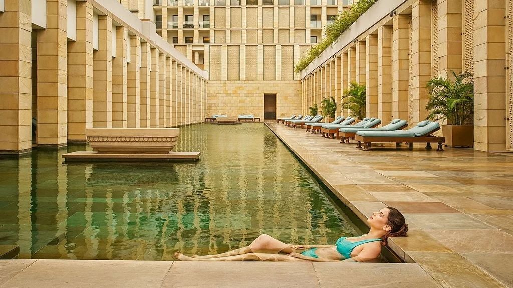 Contemporary luxury hotel exterior with stone facade, reflecting pool, and manicured gardens surrounded by tropical landscaping in New Delhi