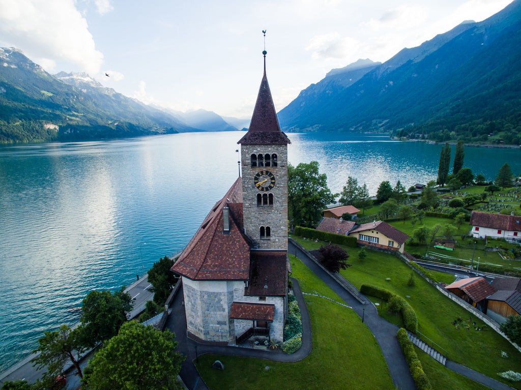 Snow-capped Alpine mountains tower over Interlaken's pristine lake, framed by lush forests and luxury chalets in foreground at sunset