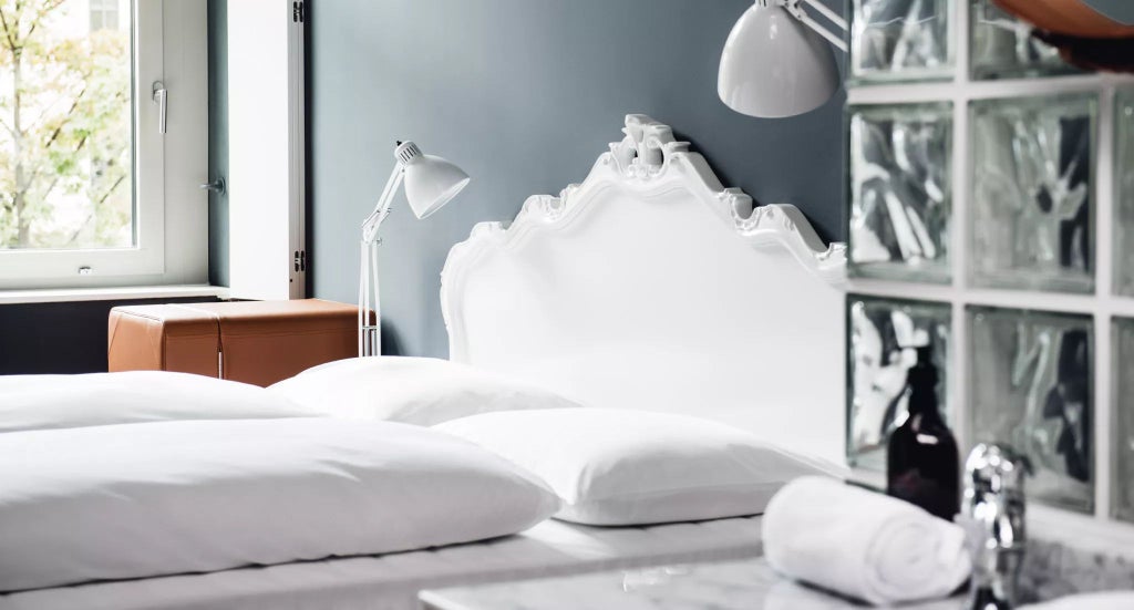 Elegant standard room at Grand Ferdinand hotel with plush white bed, wooden floors, minimalist decor, and large windows overlooking Vienna's cityscape