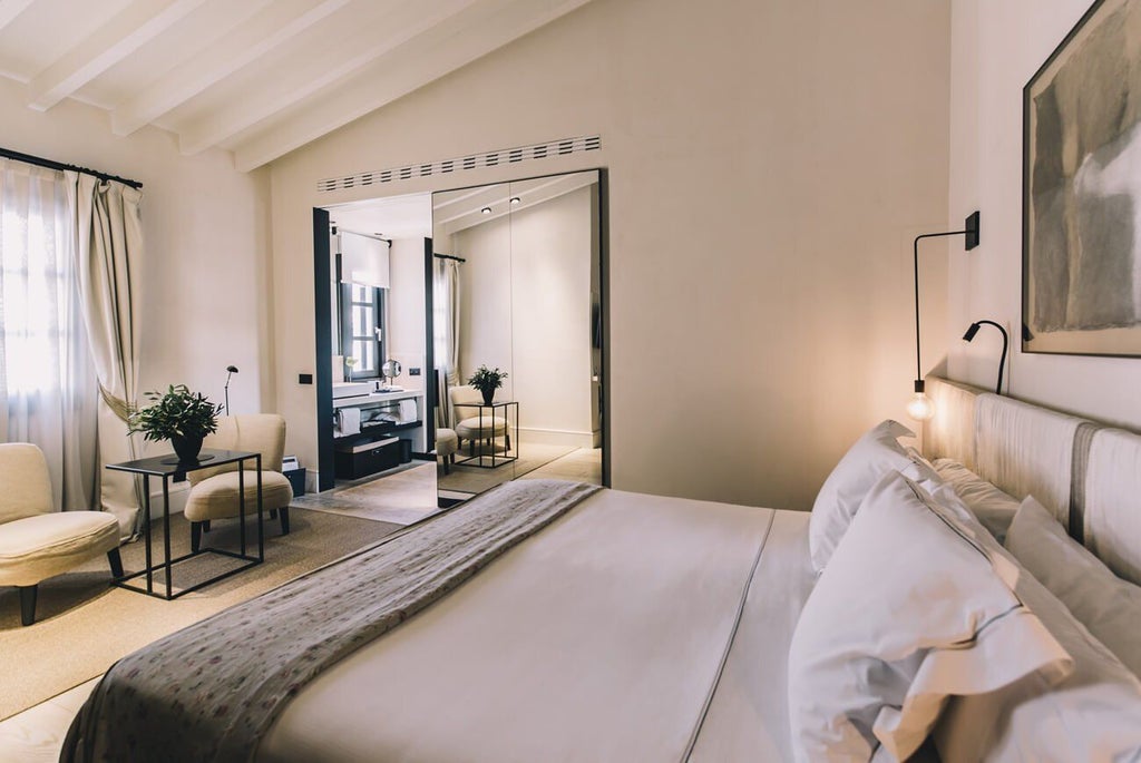 Elegant boutique hotel room with minimalist design, soft neutral tones, modern furnishings, and large windows overlooking historic Spanish architecture