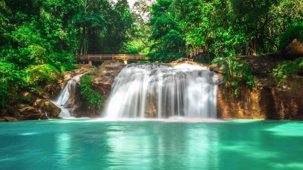 Lush tropical rainforest waterfall cascading over moss-covered rocks, with elegant picnic setup featuring white linen and gourmet cuisine amid verdant scenery of Thailand