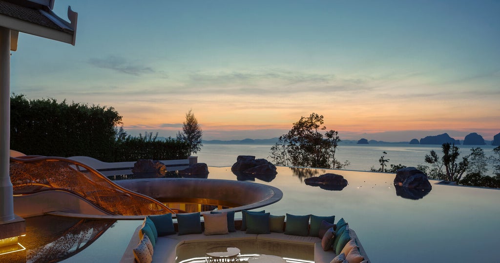 Infinity pool overlooking Andaman Sea at sunset, with private villas cascading down tropical cliffside and palm trees framing view