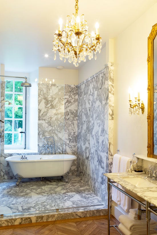 Opulent French chateau suite with ornate gold-framed mirrors, crystal chandelier, marble fireplace, and period furniture in soft white tones