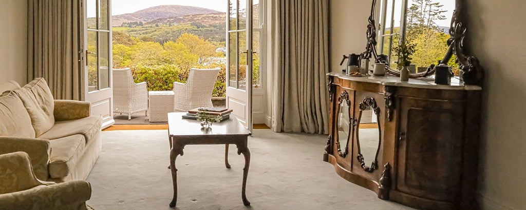 Luxurious stone-facade hotel nestled in lush Irish countryside, with elegant windows and manicured gardens against a misty mountain backdrop