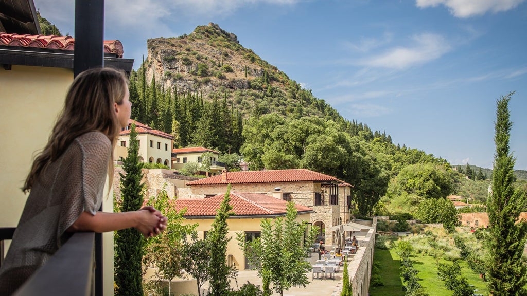 Luxurious Greek wellness resort with stone walls nestled in mountainside, featuring infinity pool overlooking lush valley at sunset