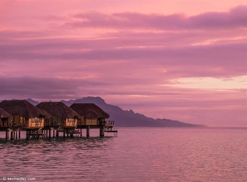 Luxurious overwater bungalows with thatched roofs stretching into turquoise Polynesian lagoon, backed by lush mountains and palm trees