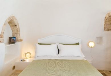 Rustic stone trullo room with white-draped bed, traditional Pugliese architecture, warm wooden accents, and elegant minimalist design in soft neutral tones