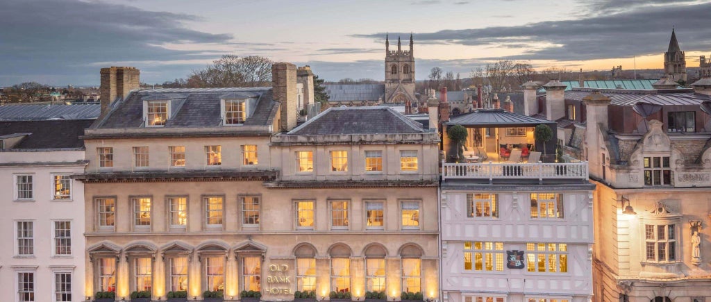 Elegant room with large windows overlooking historic cityscape, featuring classic British decor, plush furnishings, and warm ambient lighting from heritage hotel