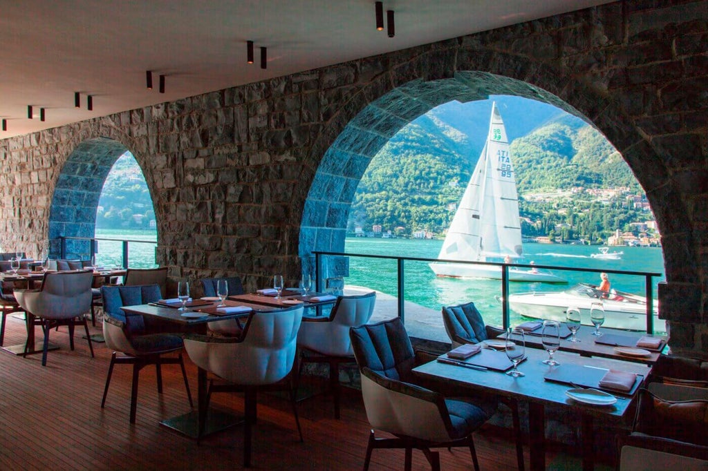 Modern minimalist hotel building with glass facade perched on Lake Como's edge, surrounded by mountains and Mediterranean gardens