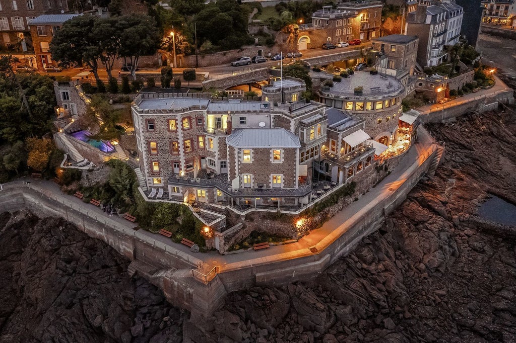 Luxurious coastal hotel with blue shutters and white facade, overlooking rocky Brittany coastline with turquoise waters and elegant architectural details