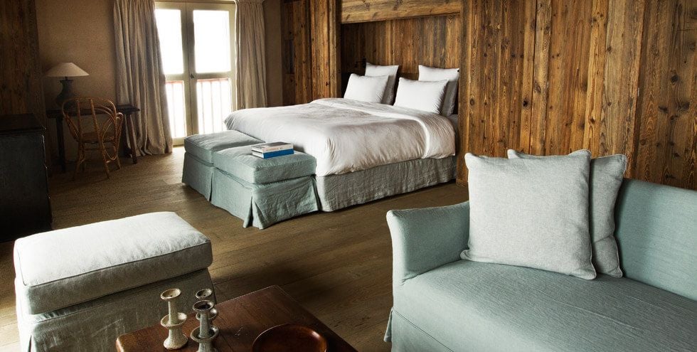 Elegant wood-paneled junior suite with plush king bed, rustic alpine decor, large windows overlooking snow-capped mountain landscape in French Alps