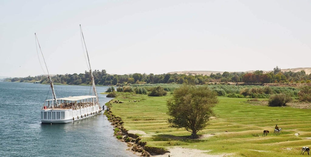 Traditional wooden dahabiya boat with elegant cream canopy sailing on the Nile, featuring ornate railings and comfortable lounge chairs on deck