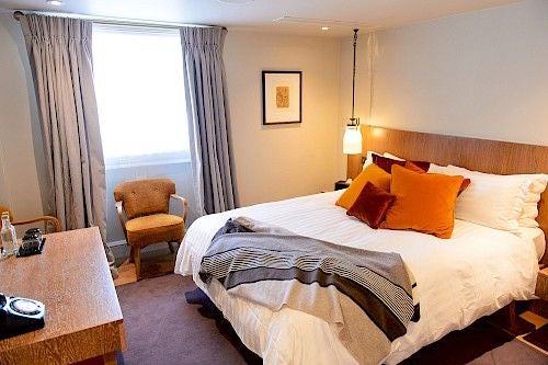 Elegant riverside hotel room with plush king bed, large windows overlooking scenic river, classic British decor in soft neutrals and warm wood tones