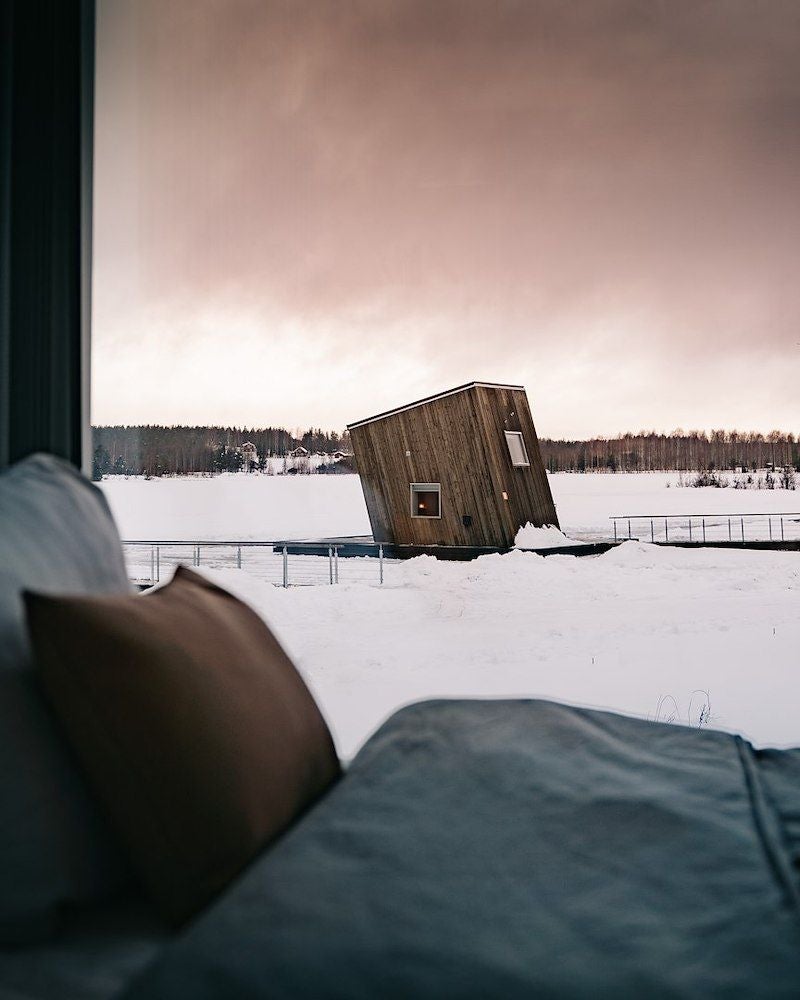 Minimalist wooden water cabin perched above frozen river, featuring floor-to-ceiling windows with stunning Nordic landscape backdrop at Arctic Bath resort