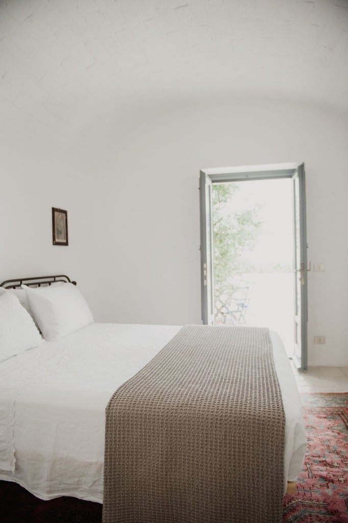 Minimalist white stone walls, rustic wooden bed, and soft linen textiles create a serene, luxurious bedroom at Masseria Moroseta in rural Puglia, Italy