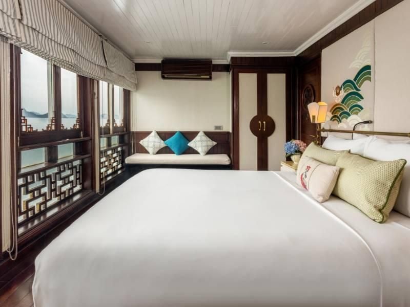 Luxurious Vietnamese cruise ship cabin with elegant wood paneling, plush king-sized bed, and panoramic window overlooking emerald Halong Bay waters