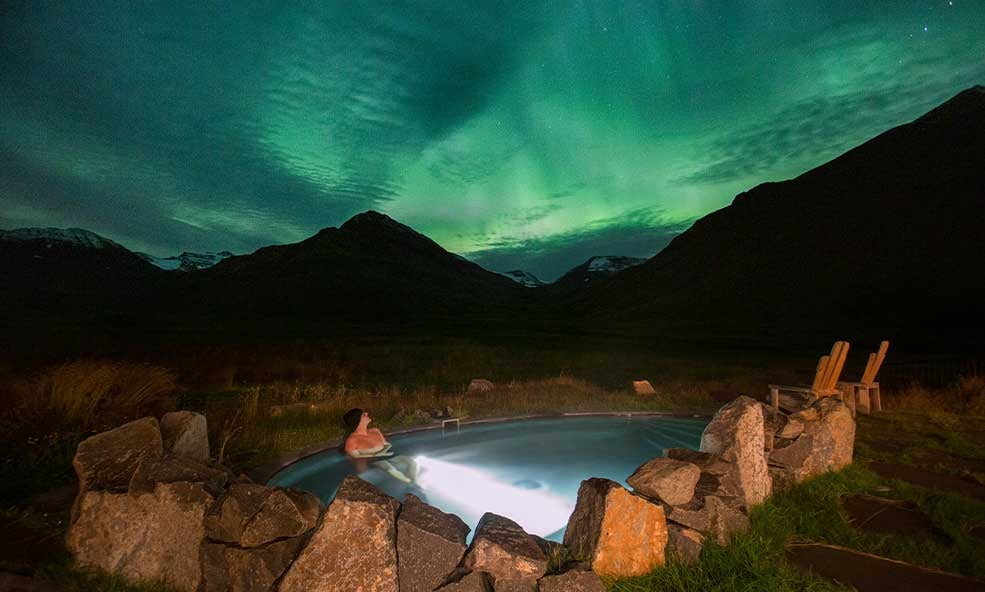 Shimmering emerald and violet auroras dance above a snow-covered Icelandic landscape, with a luxurious glass-domed accommodation nestled in the foreground