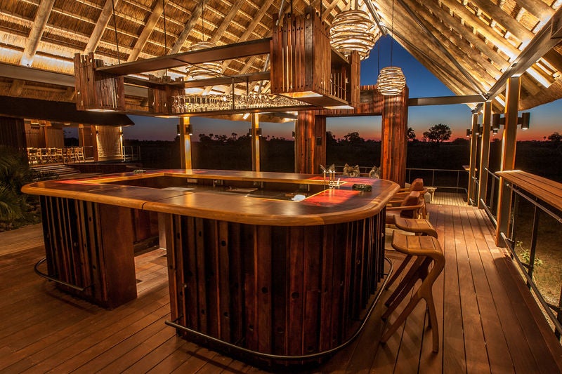 Elegant safari lodge with thatched-roof rooms elevated on wooden platforms, overlooking a pristine savannah and waterhole in Botswana