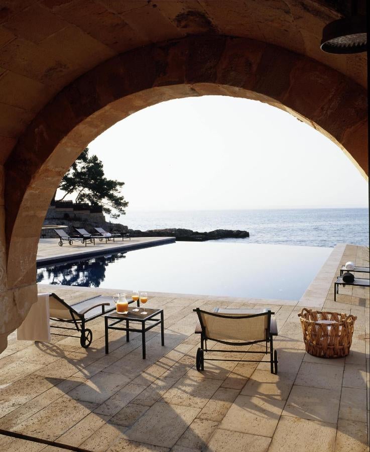 Elegant Mediterranean hotel with stone arches and terraces overlooking a turquoise sea, featuring luxurious poolside loungers at sunset