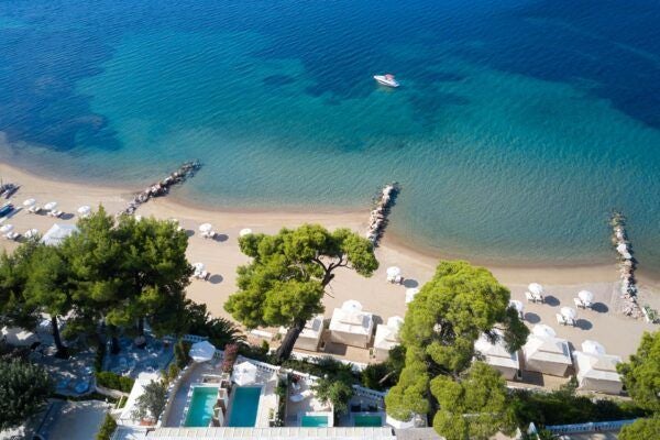 Luxurious beachfront resort with white infinity pool overlooking azure Aegean waters, surrounded by lush Mediterranean greenery and elegant stone architecture