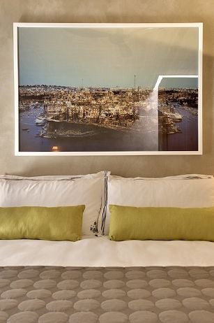 Elegant boutique hotel suite with modern minimalist design, soft neutral tones, plush king bed, and arched window overlooking historic Maltese architecture