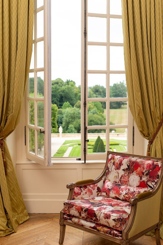 Elegant French château hotel room with ornate period furnishings, crystal chandelier, and floor-to-ceiling windows overlooking manicured gardens