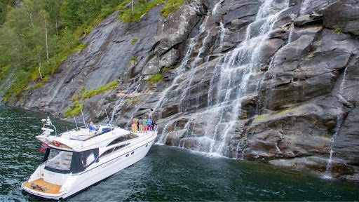 Sleek tour boat glides through dramatic Norwegian fjord, surrounded by towering cliffs and pristine waterfalls at Mostraumen